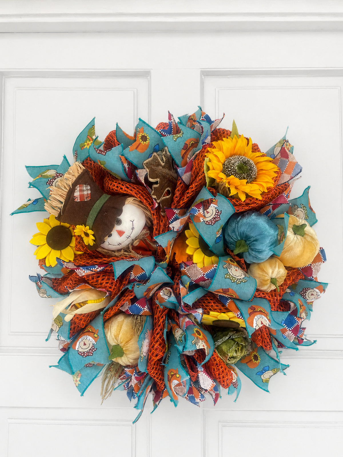 An autumn-themed fabric mesh wreath hanging on a white paneled door. The wreath features turquoise blue ribbon, orange mesh, faux pumpkins, yellow sunflowers, a small plush snowman wearing a hat, and fall patterned accents.
